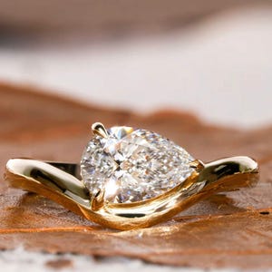 May include: A gold ring with a large, pear-cut diamond. The diamond is multi-faceted and set in a smooth, curved gold band. The ring is displayed on a brown surface, enhancing its sparkle and shine.