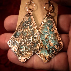 May include: A pair of handcrafted copper earrings with a textured, diamond-like shape. The earrings feature a hammered design with turquoise-colored accents, and are suspended from copper ear wires. The earrings are displayed against a neutral background.