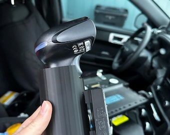 May include: A black handheld scanner with a grey top and a black cylindrical holder. The holder has the text "SF Design" on the side. The scanner is held inside a vehicle.