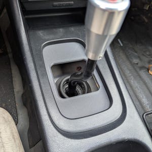 Puede incluir: Un pomo de palanca de cambios de coche plateado y negro con un logotipo rojo y blanco. El pomo está instalado en una consola de plástico gris. La consola tiene un botón y un portavasos.