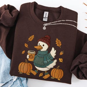 May include: A brown sweatshirt featuring a cartoon duck wearing a knit hat and scarf, holding a coffee cup, and walking past pumpkins and autumn leaves. The sweatshirt is paired with blue jeans and a silver necklace.