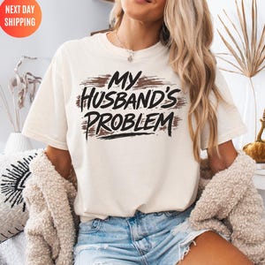 May include: Ivory-colored t-shirt with the text "MY Husband's PROBLEM" in a distressed, brushstroke design. The shirt is a casual style, suitable for everyday wear. The text is in black and brown.