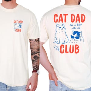 May include: Cream-colored t-shirt with "CAT DAD CLUB" in red text and cat illustrations in blue. The front features a small design, while the back displays a larger graphic with the phrase "life is better with cats."