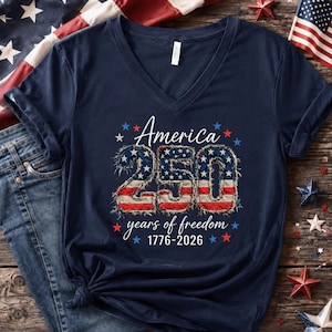 May include: Navy blue V-neck t-shirt with the text "America 250 years of freedom 1776-2026". The numbers "250" are designed with a red, white, and blue American flag pattern. The shirt is decorated with stars.