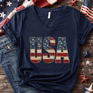 Könnte beinhalten: Marineblaues V-Ausschnitt-T-Shirt mit dem Wort "USA" in einem verwitterten Design der amerikanischen Flagge. Das T-Shirt wird auf einer rustikalen Holzoberfläche mit patriotischen Dekorationen präsentiert, darunter Sterne und eine gefaltete amerikanische Flagge.