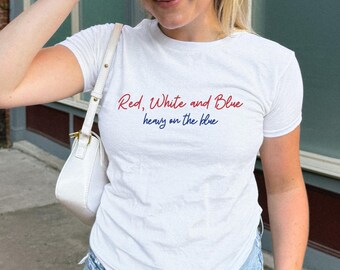 May include: White t-shirt with the text "Red, White and Blue heavy on the blue" in red and blue script. The shirt is paired with light blue denim jeans and a white baseball cap.