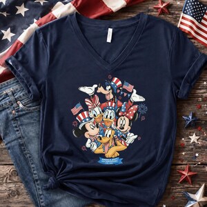 Może przedstawiać: Granatowa koszulka z dekoltem w serek z grafiką postaci Disneya: Myszki Miki, Myszki Minnie, Kaczora Donalda, Daisy, Goofy'ego i Pluto, wszyscy w patriotycznych strojach. Projekt zawiera amerykańskie flagi i fajerwerki.