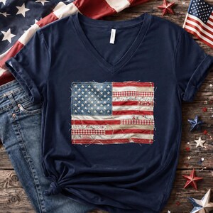May include: A navy blue V-neck t-shirt with a patchwork American flag design. The flag incorporates denim, floral, and gingham fabrics. The shirt has rolled sleeves and a knotted hem, with a patriotic theme.