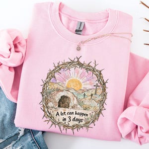 May include: A pale pink sweatshirt featuring a graphic of a sunrise over a landscape with a tomb and crosses, encircled by a thorny wreath. The text "A lot can happen in 3 days" is below the graphic. A delicate necklace is visible.
