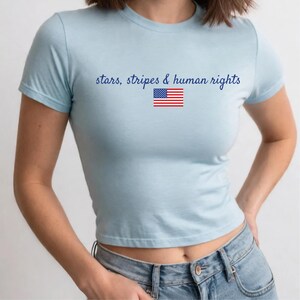 K&ouml;nnte beinhalten: Hellblaues Crop-Top mit dem Text "stars, stripes & human rights" und einer kleinen amerikanischen Flagge. Das Oberteil hat kurze &Auml;rmel und wird mit einer Jeans getragen. Das Shirt ist aus weichem Material.