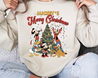 May include: Cream-colored sweatshirt featuring a festive graphic of Mickey Mouse, Minnie Mouse, Donald Duck, Goofy, and Pluto decorating a Christmas tree. The text "Mickey's Merry Christmas" is displayed above the scene in red and yellow.