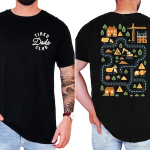 May include: Black crew neck t-shirt with the text "Tired Dads Club" in a white, circular design on the front. The back features a colorful construction site graphic with roads, houses, and construction vehicles.