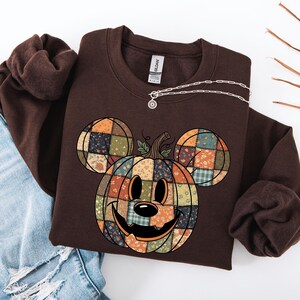 Könnte beinhalten: Ein braunes Sweatshirt mit einem Patchwork-Kürbis-Design mit Mickey Mouse-Ohren. Der Kürbis hat ein lächelndes Gesicht und besteht aus verschiedenen Stoffmustern in Orange, Grün und Blau. Eine silberne Halskette mit einem kleinen Anhänger ist auf dem Sweatshirt drapiert.