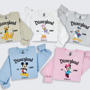 Puede incluir: Una colecci&oacute;n de sudaderas de Disneyland Tokio en varios colores: beige, blanco, gris, azul claro y rosa. Cada sudadera presenta un personaje diferente de Disney, el logotipo de Disneyland, el a&ntilde;o 1983 y la palabra "TOKYO".