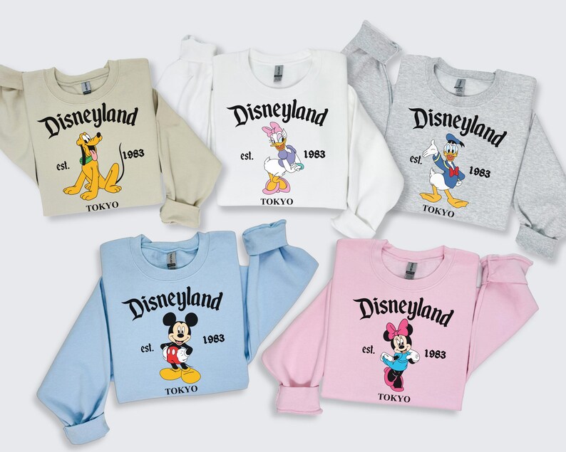 Puede incluir: Una colecci&oacute;n de sudaderas de Disneyland Tokio en varios colores: beige, blanco, gris, azul claro y rosa. Cada sudadera presenta un personaje diferente de Disney, el logotipo de Disneyland, el a&ntilde;o 1983 y la palabra "TOKYO".