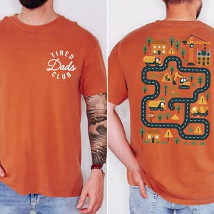 May include: Burnt orange t-shirt with "Tired Dads Club" text on the front. The back features a construction-themed graphic with roads, buildings, and construction vehicles. A fun, casual tee for dads.