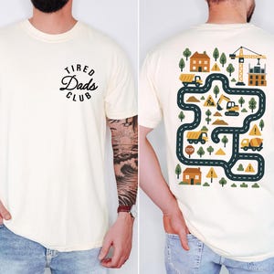May include: Cream-colored t-shirt with the text "Tired Dads Club" on the front. The back features a colorful construction-themed graphic with roads, trucks, trees, and a crane. A fun and playful design for dads.
