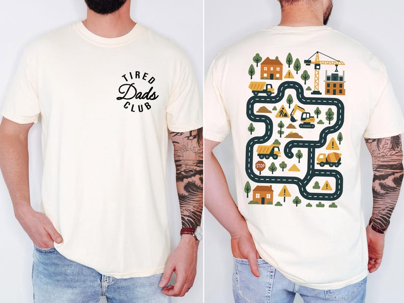May include: Cream-colored t-shirt with the text "Tired Dads Club" on the front. The back features a colorful construction-themed graphic with roads, trucks, trees, and a crane. A fun and playful design for dads.