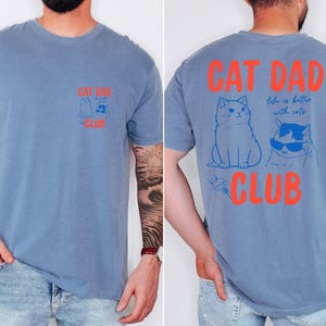 May include: Blue short-sleeved t-shirt with the text "CAT DAD CLUB" in orange. The front features a cat graphic, and the back has two cat illustrations and the phrase "life is better with cats."