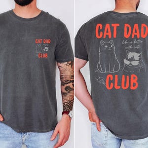 May include: Dark grey t-shirt with "CAT DAD CLUB" in red text. The front features a small graphic of two cats, and the back displays a larger design with two cats and the phrase "life is better with cats."