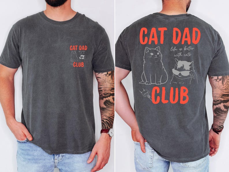 May include: Dark grey t-shirt with "CAT DAD CLUB" in red text. The front features a small graphic of two cats, and the back displays a larger design with two cats and the phrase "life is better with cats."