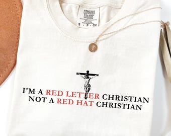 May include: Cream-colored t-shirt with the text "I'M A RED LETTER CHRISTIAN NOT A RED HAT CHRISTIAN" in black and red. A gold necklace with a pendant is draped across the shirt. The shirt has a crew neck and short sleeves.