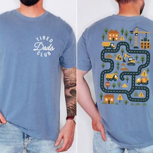 May include: Blue short-sleeved t-shirt with the text "Tired Dads Club" on the front. The back features a colorful construction-themed graphic with roads, trucks, and a crane. The shirt is made of a soft material.
