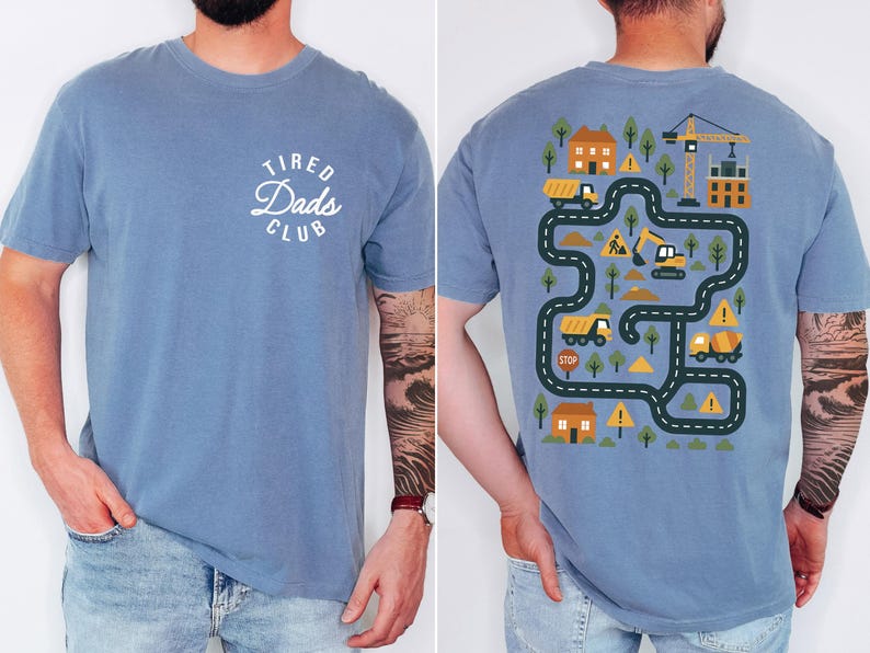 May include: Blue short-sleeved t-shirt with the text "Tired Dads Club" on the front. The back features a colorful construction-themed graphic with roads, trucks, and a crane. The shirt is made of a soft material.