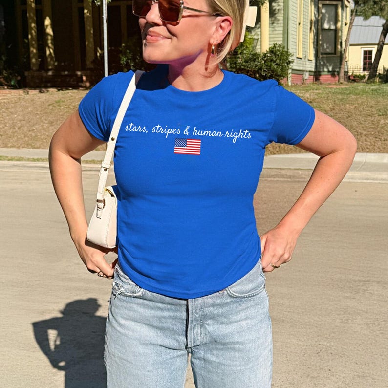 K&ouml;nnte beinhalten: K&ouml;nigsblaues T-Shirt mit dem Text "stars, stripes & human rights" und einer kleinen amerikanischen Flagge. Das Shirt wird mit hellblauen Jeans kombiniert. Der Look ist l&auml;ssig und patriotisch.