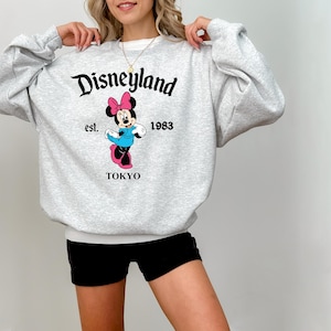 Puede incluir: Una sudadera gris claro con la palabra "Disneyland" en escritura negra. Debajo del texto hay un gr&aacute;fico de Minnie Mouse con un lazo rosa, un vestido azul y zapatos rosas. La sudadera tambi&eacute;n tiene el texto "est. 1983 TOKYO".