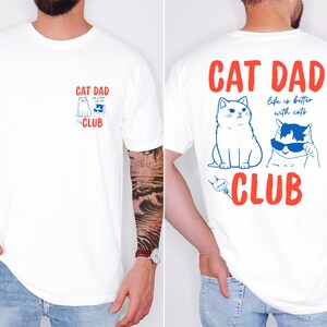 May include: White t-shirt with a red and blue graphic. The front features a small design with two cats and the words "CAT DAD CLUB". The back has a larger design with two cats, the words "CAT DAD", and "life is better with cats".