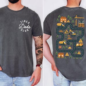 May include: Dark gray t-shirt with the text "Tired Dads Club" in a white, circular design on the front. The back features a colorful construction-themed graphic with roads, vehicles, and buildings.