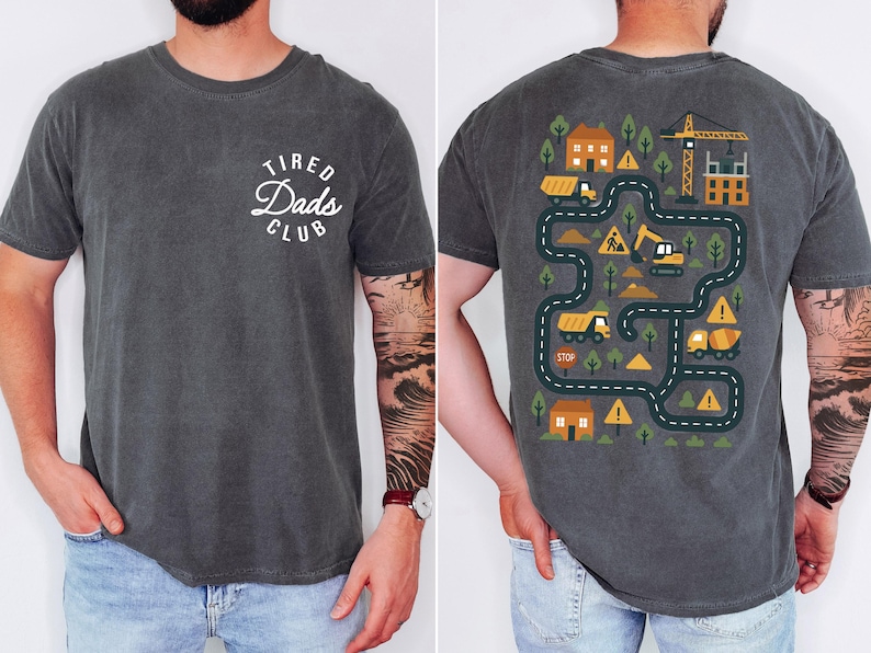 May include: Dark gray t-shirt with the text "Tired Dads Club" in a white, circular design on the front. The back features a colorful construction-themed graphic with roads, vehicles, and buildings.