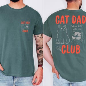 May include: A teal-colored t-shirt with the text "CAT DAD CLUB" in red. The front of the shirt has a small graphic of a cat and the text "CAT DAD CLUB". The back of the shirt has a larger graphic of two cats and the text "Life is better with cats".