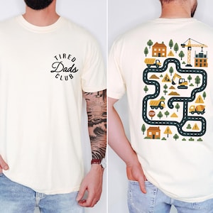 May include: Cream-colored t-shirt with the text "Tired Dads Club" in a black, circular design on the front. The back features a colorful construction-themed graphic with trucks, houses, trees, and a crane.