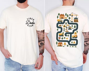 May include: Cream-colored t-shirt with the text "Tired Dads Club" in a black, circular design on the front. The back features a colorful construction-themed graphic with trucks, houses, trees, and a crane.