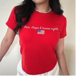 K&ouml;nnte beinhalten: Ein rotes T-Shirt mit dem wei&szlig;en Schriftzug "stars, stripes & human rights" und einer kleinen amerikanischen Flagge. Das Shirt hat kurze &Auml;rmel und einen Rundhalsausschnitt.