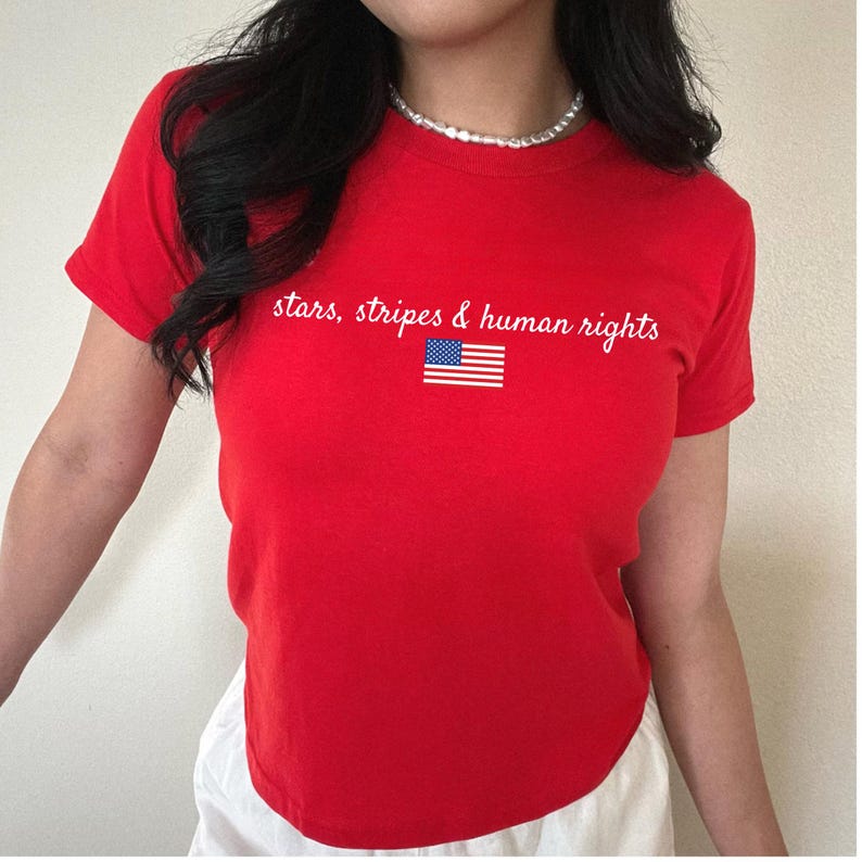 K&ouml;nnte beinhalten: Ein rotes T-Shirt mit dem wei&szlig;en Schriftzug "stars, stripes & human rights" und einer kleinen amerikanischen Flagge. Das Shirt hat kurze &Auml;rmel und einen Rundhalsausschnitt.