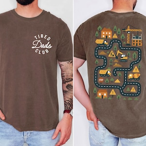 May include: Brown t-shirt with "Tired Dads Club" text on the front. The back features a colorful construction-themed graphic with roads, houses, and construction vehicles. The shirt is made of a soft material.