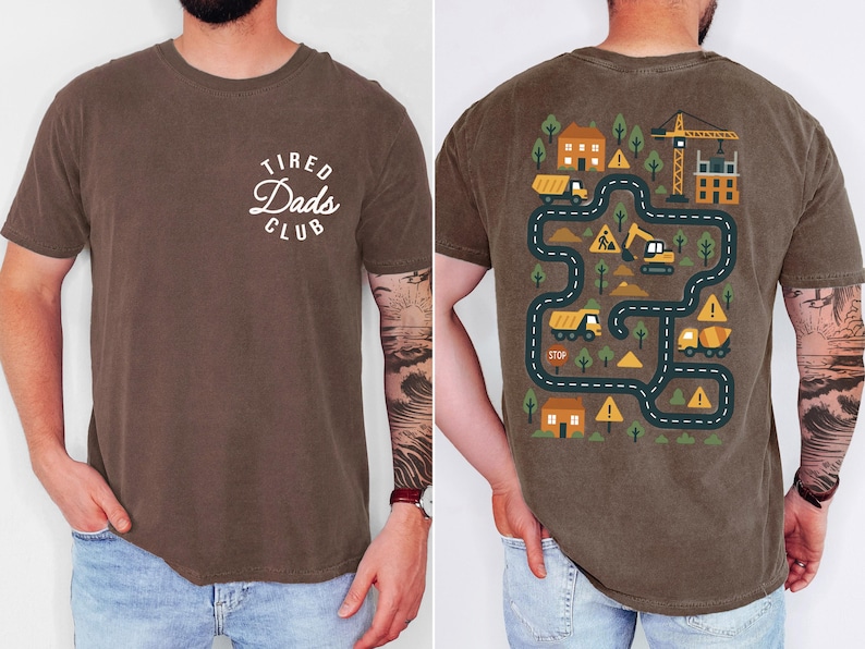 May include: Brown t-shirt with "Tired Dads Club" text on the front. The back features a colorful construction-themed graphic with roads, houses, and construction vehicles. The shirt is made of a soft material.