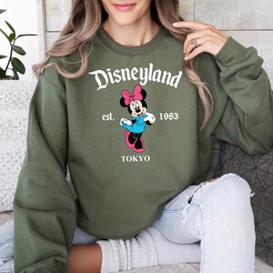 Puede incluir: Sudadera verde oliva con un dise&ntilde;o de Minnie Mouse con lazo rosa, vestido azul y zapatos rosas. El texto "Disneyland est. 1983 TOKYO" est&aacute; impreso encima y debajo del dise&ntilde;o. La sudadera es de estilo casual.
