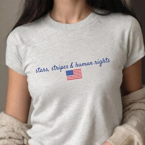 K&ouml;nnte beinhalten: Hellgraues T-Shirt mit dem blauen Schriftzug "stars, stripes & human rights". Darunter befindet sich eine kleine amerikanische Flagge. Das Shirt hat kurze &Auml;rmel.