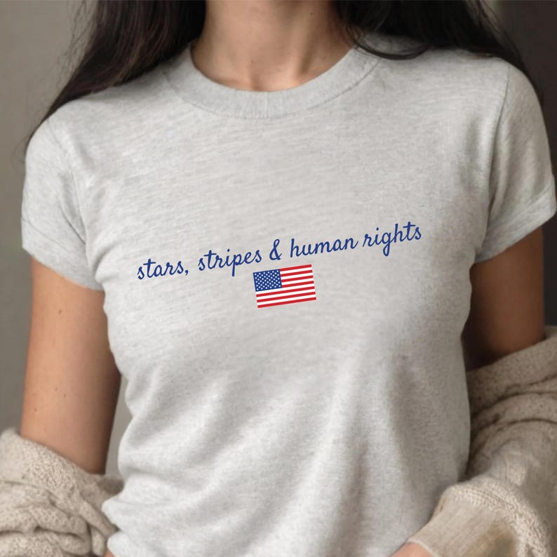K&ouml;nnte beinhalten: Hellgraues T-Shirt mit dem blauen Schriftzug "stars, stripes & human rights". Darunter befindet sich eine kleine amerikanische Flagge. Das Shirt hat kurze &Auml;rmel.