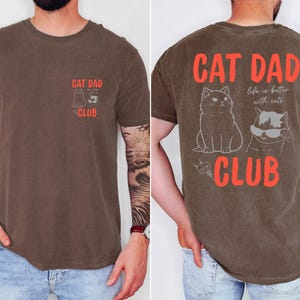 May include: Brown t-shirt with the text "CAT DAD CLUB" in red. The front of the shirt has a small graphic of a cat and the text "CAT DAD CLUB". The back of the shirt features two cat illustrations and the phrase "life is better with cats".