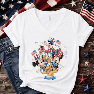 Może przedstawiać: Biała koszulka z dekoltem w serek z grafiką postaci Disneya: Myszki Miki, Myszki Minnie, Kaczora Donalda, Kaczki Daisy, Goofy'ego i Pluto, wszyscy w patriotycznych strojach. Projekt zawiera amerykańskie flagi i fajerwerki. Niebieski baner głosi "America 250th Anniversary".