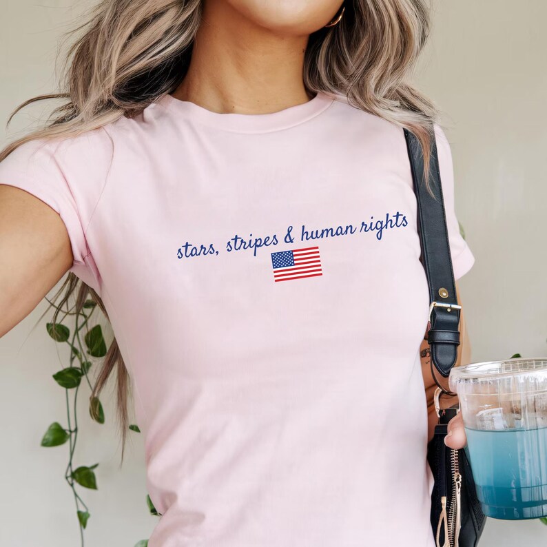 K&ouml;nnte beinhalten: Hellrosa T-Shirt mit blauem Schriftzug "stars, stripes & human rights". Darunter befindet sich eine kleine amerikanische Flagge. Das T-Shirt hat kurze &Auml;rmel.