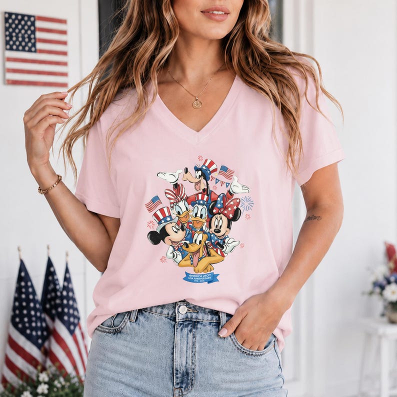 Może przedstawiać: Jasnor&oacute;żowa koszulka z dekoltem w serek z grafiką Myszki Miki, Myszki Minnie, Kaczora Donalda, Goofy'ego i Pluto w patriotycznych strojach. Projekt zawiera amerykańskie flagi i fajerwerki, świętując USA.