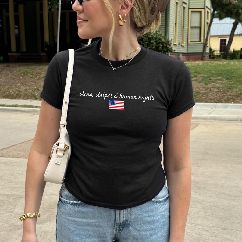 K&ouml;nnte beinhalten: Schwarzes T-Shirt mit Rundhalsausschnitt, dem Text "stars, stripes & human rights" und einer kleinen amerikanischen Flagge. Ein schlichtes, l&auml;ssiges Oberteil.