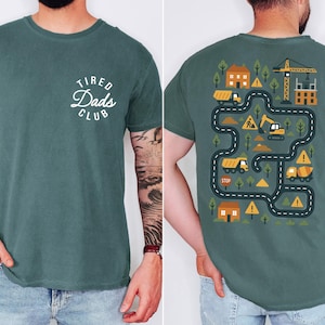 May include: A teal-colored t-shirt with the text "Tired Dads Club" in a white, circular design on the front. The back features a construction-themed graphic with trucks, a crane, and a road map design.