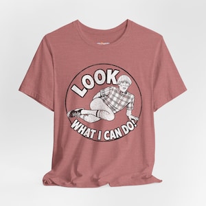 Könnte beinhalten: Staubrosafarbenes T-Shirt mit einer Schwarz-Weiß-Grafik einer Person in einem karierten Hemd und dem Text "LOOK WHAT I CAN DO!". Die Grafik befindet sich in einem schwarzen Kreis. Das T-Shirt hat kurze Ärmel und einen Rundhalsausschnitt.
